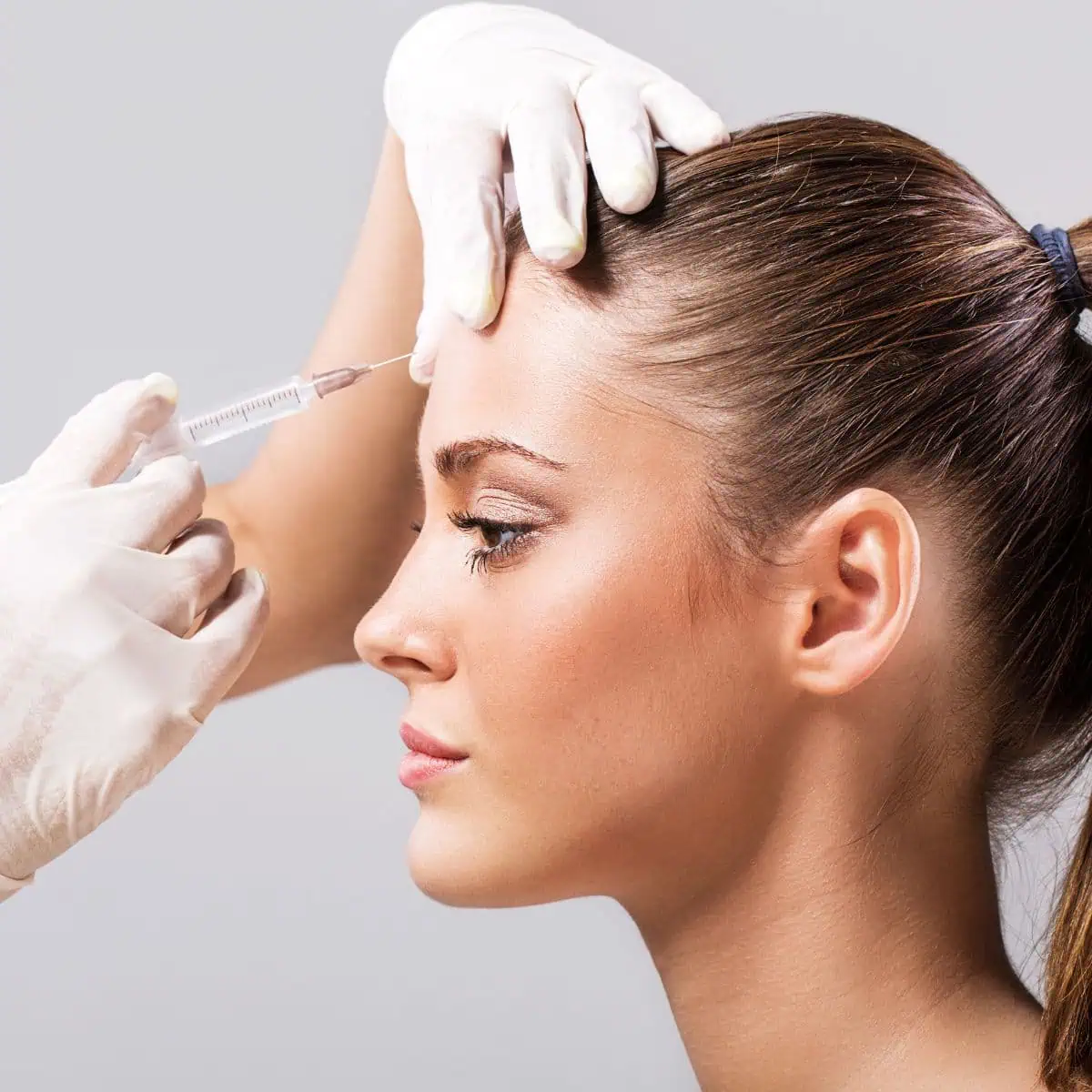 Femme de profil, cheveux attachés, recevant une injection au front près de la tempe par des mains gantées tenant une seringue sur fond neutre.