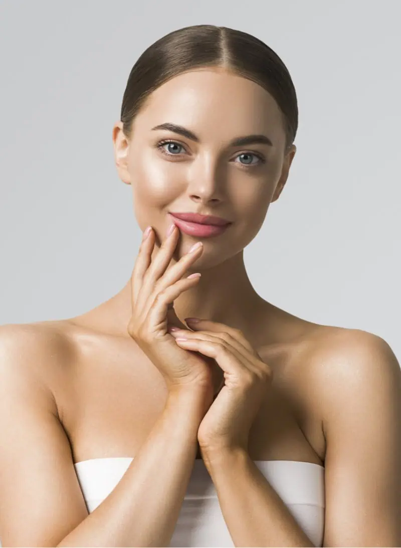 toxine botulique Jeune femme au teint lumineux et sourire doux, cheveux lissés en arrière, mains jointes près du visage et portant un haut bandeau blanc sur fond gris neutre.