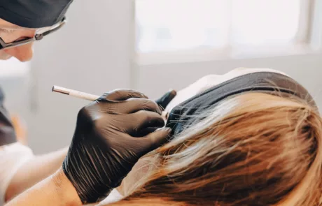 clinique esthétique des musicnes à Nice Technicien·ne en bonnet et lunettes, ganté·e de noir, utilise un stylet pour réaliser un maquillage semi-permanent des sourcils sur une cliente allongée portant un bandeau noir.