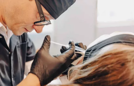 clinique esthétique des musicnes à Nice Un praticien coiffé d'une charlotte et portant des lunettes et des gants noirs administre une injection à l'aide d'une seringue près de l'œil/sourcil d'une patiente allongée dont la tête est maintenue par un bandeau.