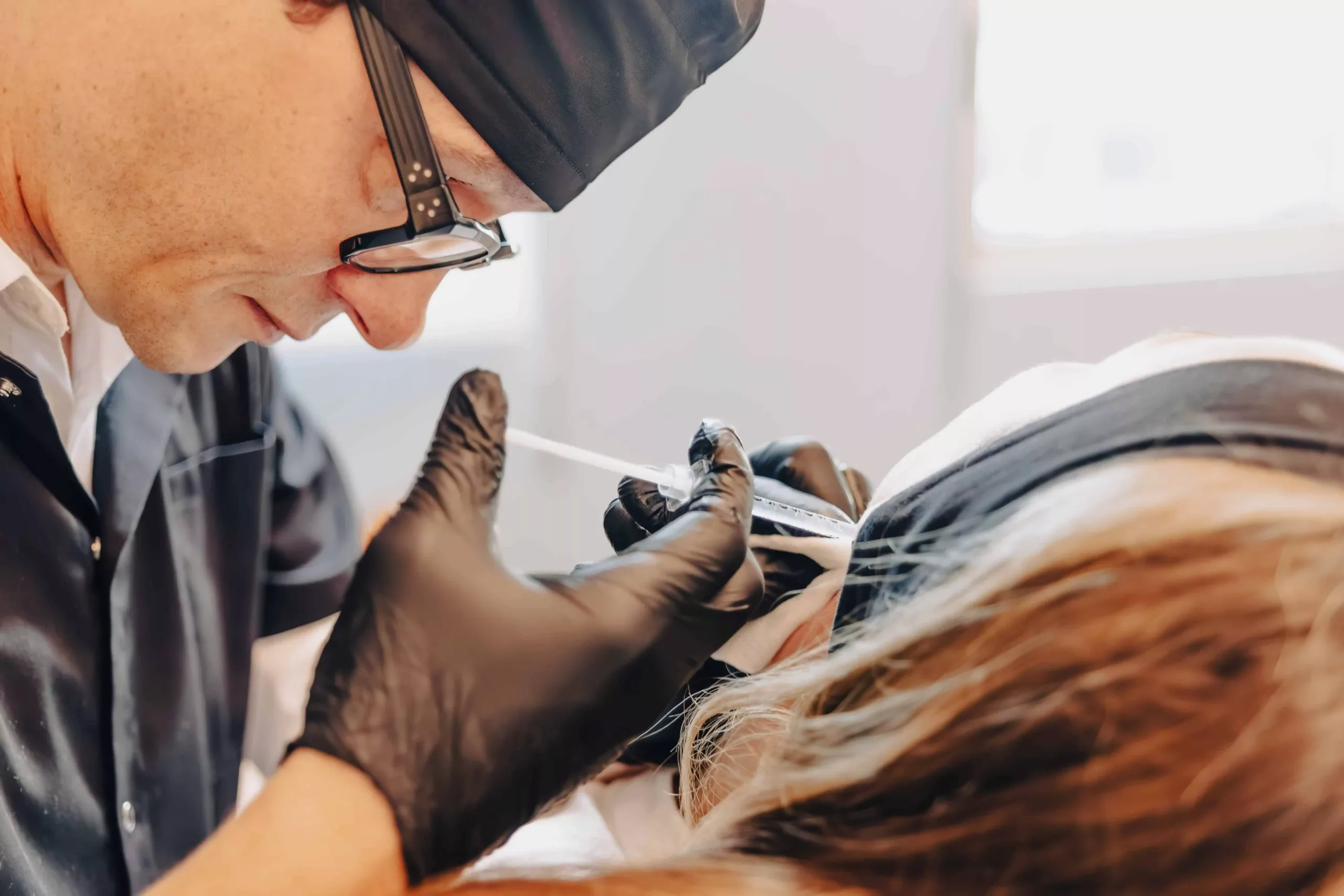 IMAGE-38 Un praticien coiffé d'une charlotte et portant des lunettes et des gants noirs administre une injection à l'aide d'une seringue près de l'œil/sourcil d'une patiente allongée dont la tête est maintenue par un bandeau.