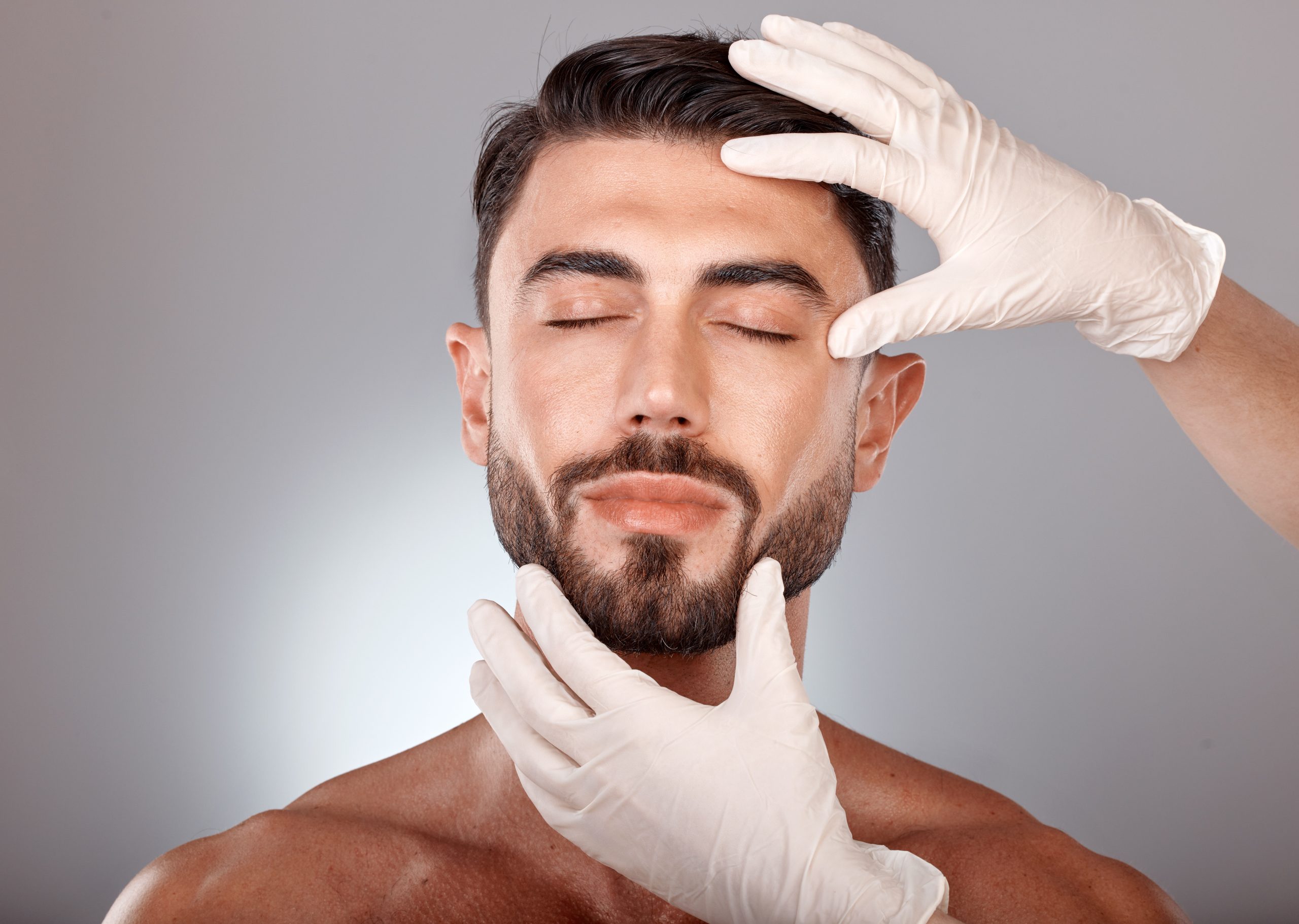 Soins esthétiques pour hommes au Centre Esthétique des Musiciens à Nice Homme torse nu aux yeux fermés et barbe soignée, le visage examiné par des mains gantées blanches tenant le front et le menton sur un fond gris neutre.