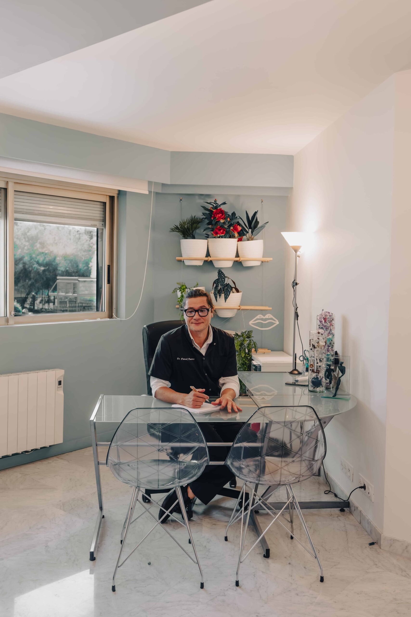 clinique des musicnes à Nice 41 Homme portant des lunettes et une blouse noire assis derrière un bureau en verre dans un bureau lumineux, écrivant sur un document, deux chaises transparentes devant lui, étagères murales avec plantes en pots et lampe sur pied à droite.