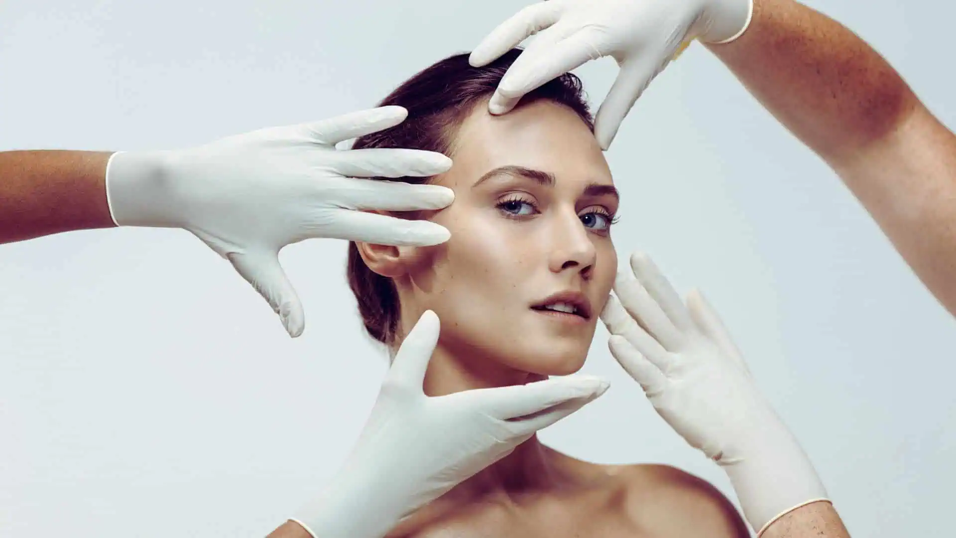 Portrait d'une femme au visage dégagé et au regard posé, entourée et examinée par plusieurs mains gantées blanches qui encadrent son visage, évoquant une consultation esthétique.