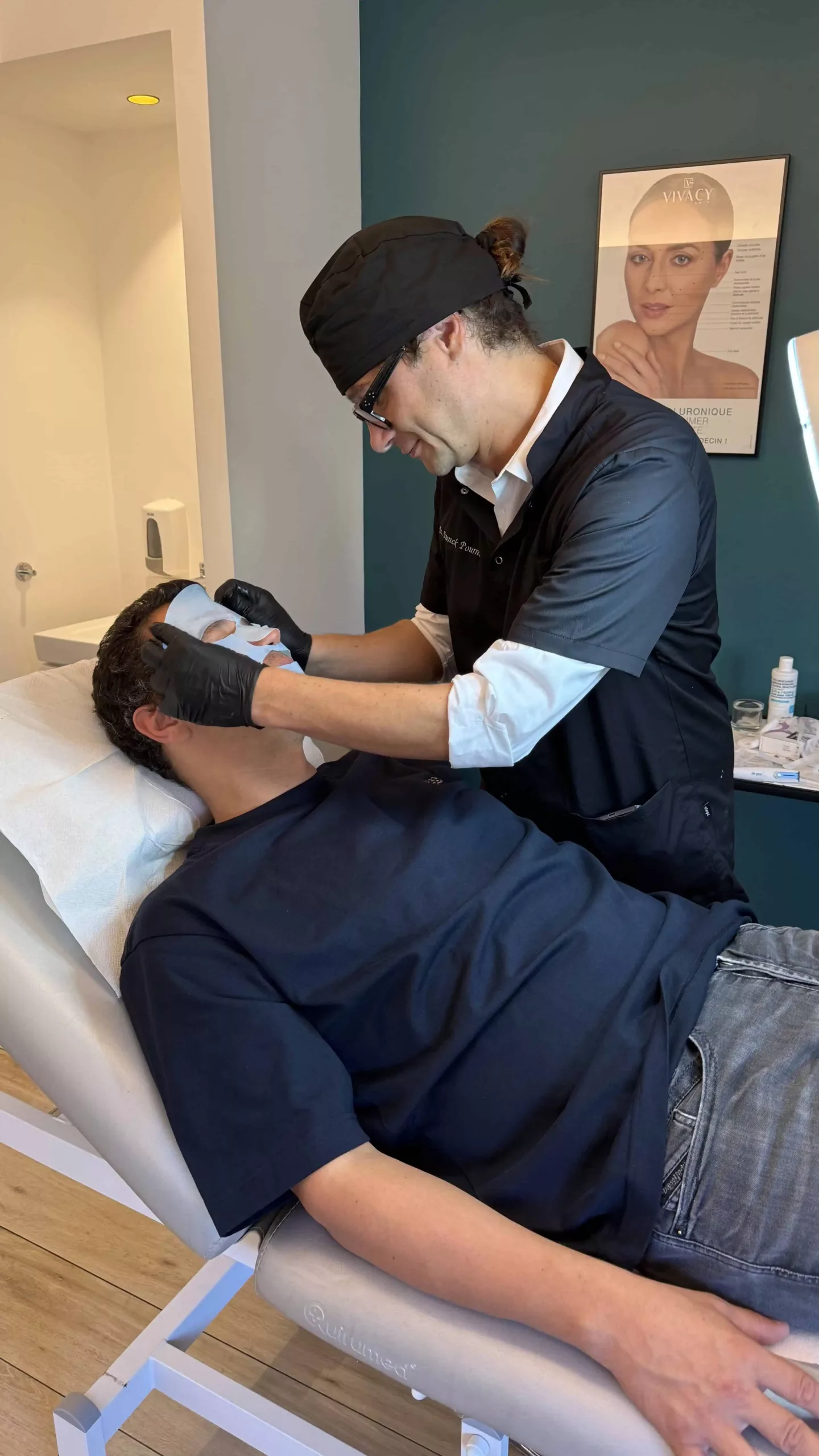 Un praticien en tenue noire et gants applique un masque facial bleu sur le visage d'un homme allongé sur une table de soin dans une salle de clinique esthétique, avec un poster de visage accroché au mur en arrière-plan.
