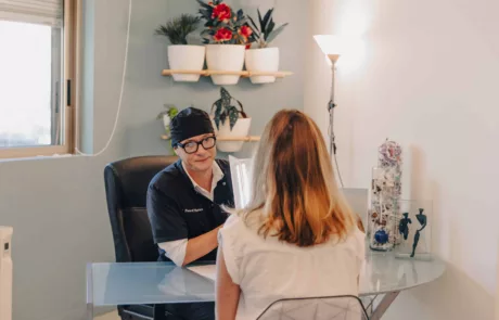 clinique esthétique des musicnes à Nice Un professionnel en blouse noire et bonnet, portant des lunettes et tenant une lampe, consulte une patiente assise de dos à un bureau dans un cabinet lumineux décoré de plantes en pots et d'une lampe sur pied.