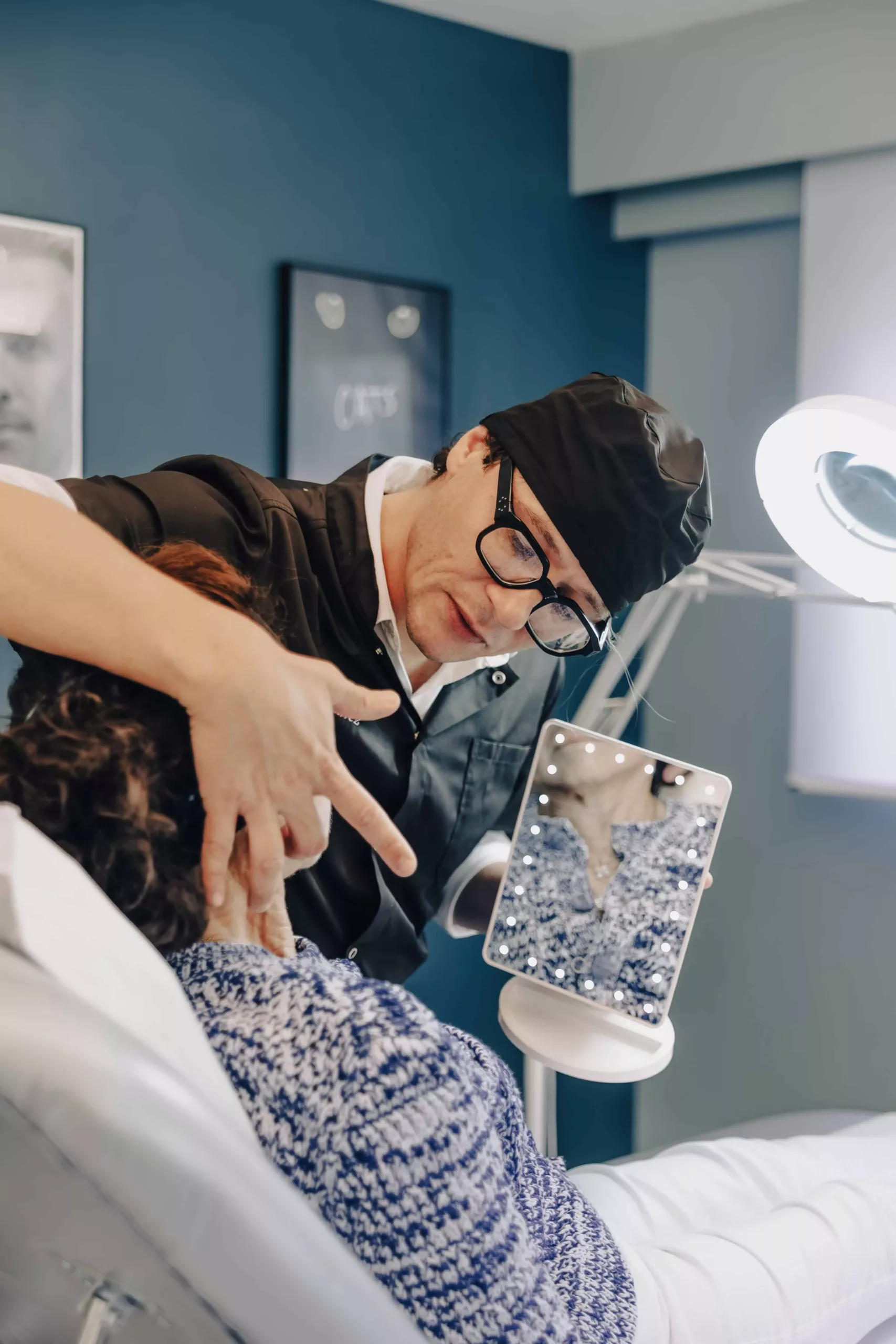 clinique des musicnes à Nice 32 Un praticien en tenue sombre et bonnet noir, portant des lunettes, examine le cou d'une patiente assise et tient un miroir entouré de LED montrant le reflet de la femme en pull bleu dans un cabinet médical.