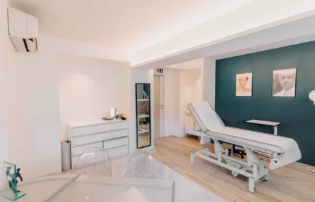 clinique des musicnes à Nice 50 Cabinet médical esthétique lumineux avec une table d'examen réglable recouverte de drap à droite, mur d'accent bleu orné de deux portraits encadrés, meuble bas blanc à tiroirs, grand miroir vertical et sol en bois clair.