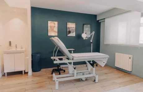 clinique des musicnes à Nice 54 Cabinet médical moderne avec une table d'examen blanche réglable au centre, un mur vert foncé décoré de trois affiches, une lampe d'examen articulée, un lavabo et une poubelle à gauche et une grande fenêtre à store à droite.