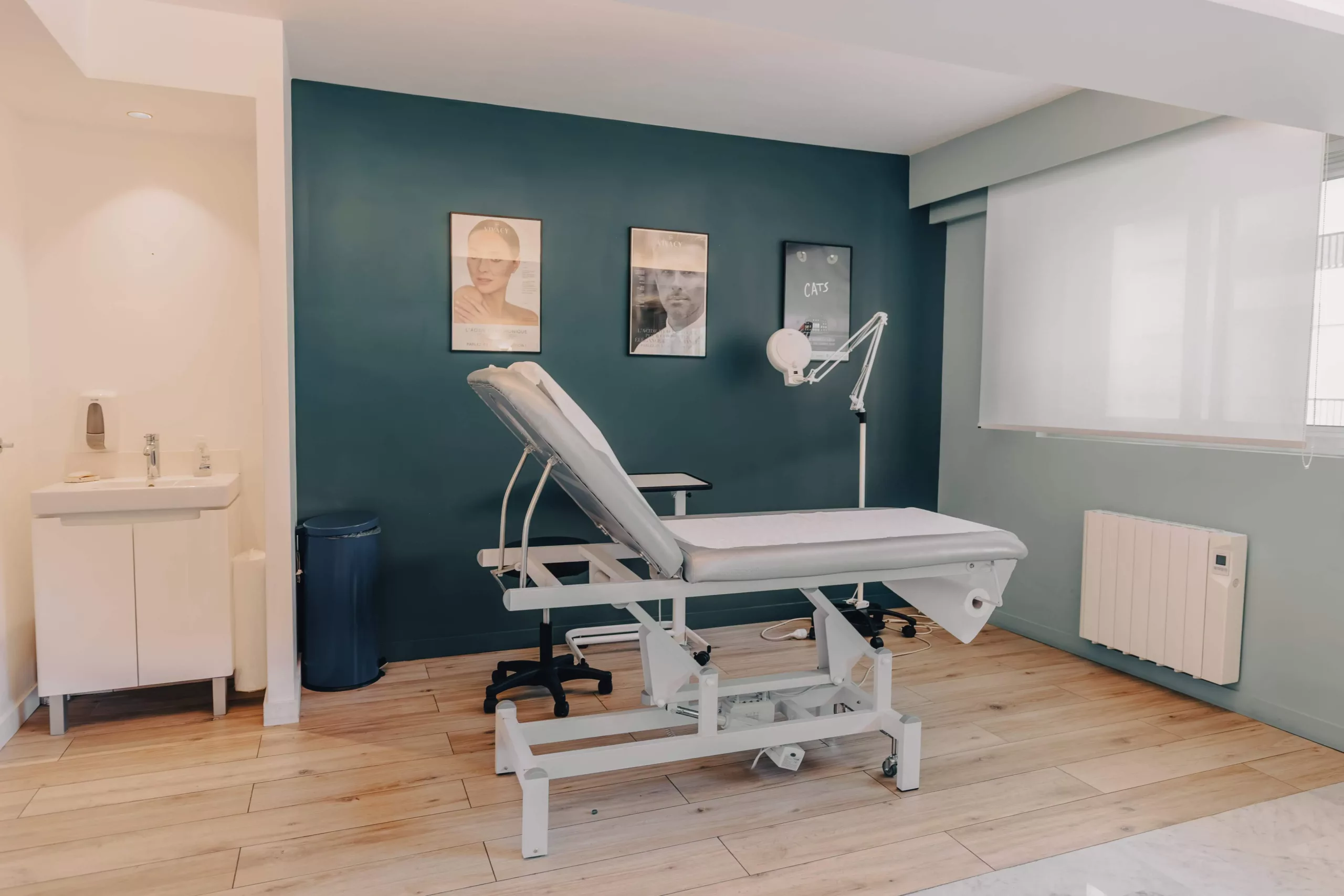 clinique des musicnes à Nice 54 Cabinet médical moderne avec une table d'examen blanche réglable au centre, un mur vert foncé décoré de trois affiches, une lampe d'examen articulée, un lavabo et une poubelle à gauche et une grande fenêtre à store à droite.